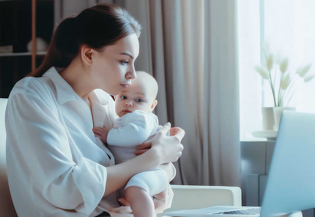 Young mother with small baby in her arms sits in front of laptop in home office